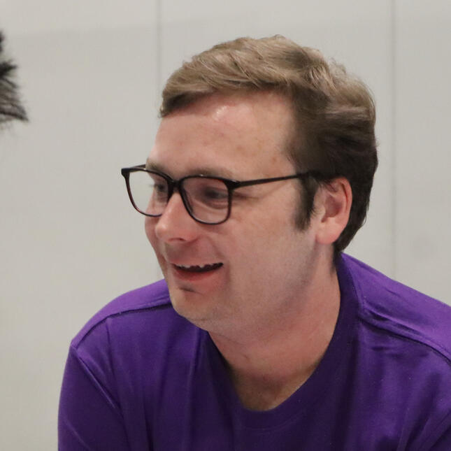 A photo of Felix Thomas, a man wearing glasses looking off to the left side of the photo, He is smiling and against a plain backdrop.